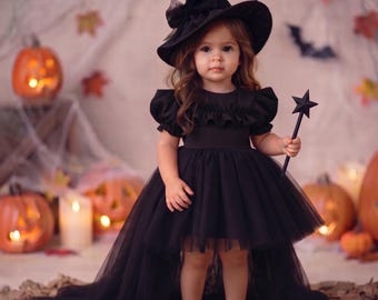 May include: A black dress with a high-low tulle skirt, ruffled bodice, and short puff sleeves. The outfit includes a matching black pointed witch hat with a decorative bow and a black star-shaped wand. The ensemble is solid black throughout.