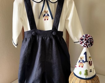 Puede incluir: Un conjunto de lino blanco con una pajarita negra y un oso bordado. El conjunto incluye una camisa blanca con cuello Peter Pan, tirantes negros y pantalones cortos de lino negro. El conjunto se combina con un gorro de fiesta de fieltro blanco con un pompón rojo, blanco y azul y detalles bordados.