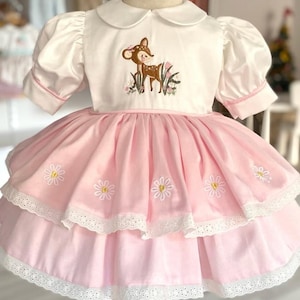 May include: A white and pink dress for a child. The top is white with a Peter Pan collar and an embroidered deer design. The skirt is layered pink with daisy embroidery and white lace trim. The dress is displayed on a mannequin.