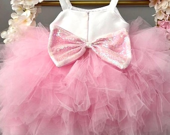 May include: A pink tulle dress with a white bodice and a large sequined bow on the back. The dress has spaghetti straps and is hanging on a white hanger. The dress is surrounded by pink and white flowers.