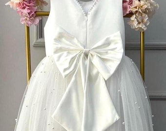 May include: A white sleeveless dress with a large satin bow on the back. The dress features a tulle skirt adorned with small pearls. The neckline is embellished with delicate beading. The dress is hanging on a white hanger, with pink and cream flowers in the background.