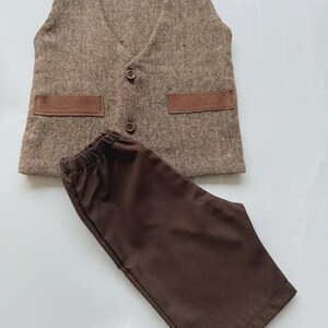 May include: A brown tweed vest with two brown pockets and two brown buttons, paired with brown pants. The vest has a V-neck design. The pants have an elastic waistband. The outfit is displayed on a white surface.