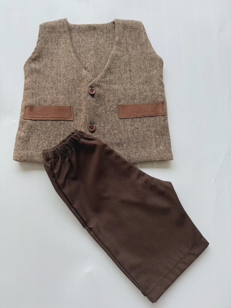 May include: A brown tweed vest with two brown pockets and two brown buttons, paired with brown pants. The vest has a V-neck design. The pants have an elastic waistband. The outfit is displayed on a white surface.