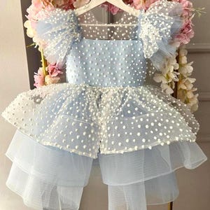 May include: A light blue dress with white polka dots and layered tulle skirt. The dress features ruffled sleeves and a square neckline. The dress is hanging on a white hanger, with pink and white flowers in the background.