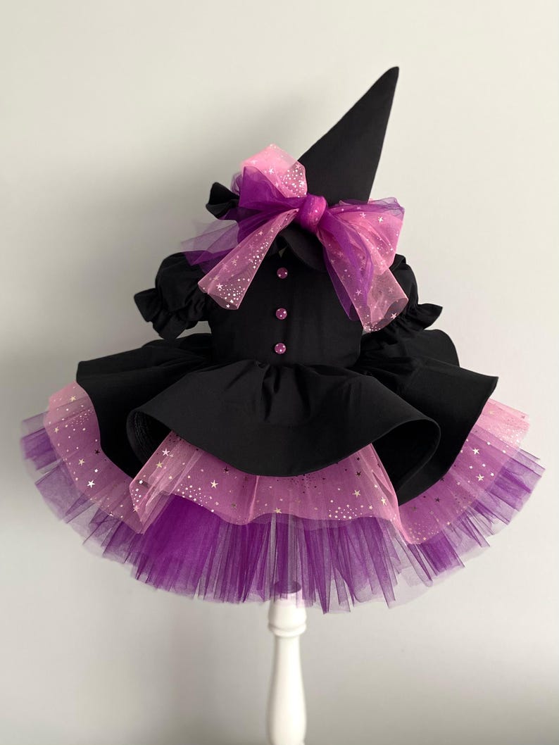May include: A black witch costume with a matching hat. The dress features a layered skirt with purple and pink tulle, adorned with star-shaped glitter. A large pink and purple bow accents the bodice.