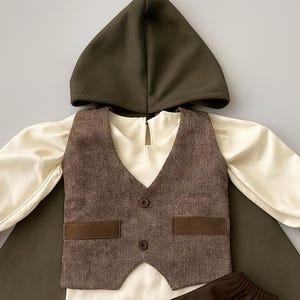 May include: A child's costume set featuring a dark olive green hooded cape, a brown tweed vest with two buttons and two pockets, and cream-colored long-sleeved shirt with ruffled cuffs. The outfit is designed for a child.
