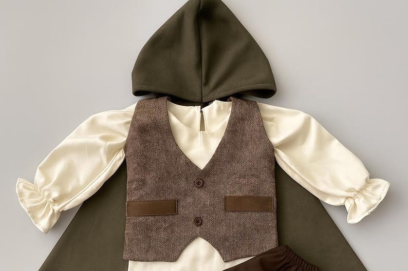May include: A child's costume set featuring a dark olive green hooded cape, a brown tweed vest with two buttons and two pockets, and cream-colored long-sleeved shirt with ruffled cuffs. The outfit is designed for a child.