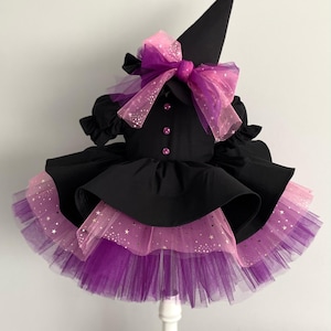 May include: A black witch costume with a matching pointed hat. The dress features a layered skirt with purple and pink tulle, adorned with star-shaped glitter. A large pink and purple bow accents the bodice.
