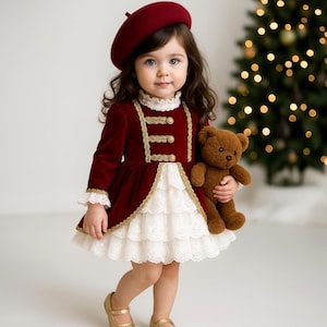May include: A young child wearing a burgundy velvet dress with gold trim, a matching beret, and gold shoes. The dress has a white lace skirt and gold buttons. The child holds a brown teddy bear. A Christmas tree is in the background.