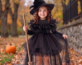 May include: A black witch costume featuring a pointed hat with feathers, a long-sleeved dress with a ruffled tulle bodice, and a layered high-low tulle skirt. The outfit is paired with a wooden broomstick and a plastic orange jack-o'-lantern bucket.