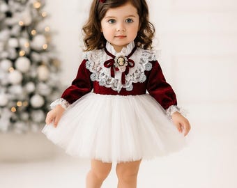 May include: A young child in a burgundy velvet dress with a white tulle skirt and lace collar. A matching burgundy bow adorns the hair. The outfit is completed with burgundy shoes. The background includes a Christmas tree.