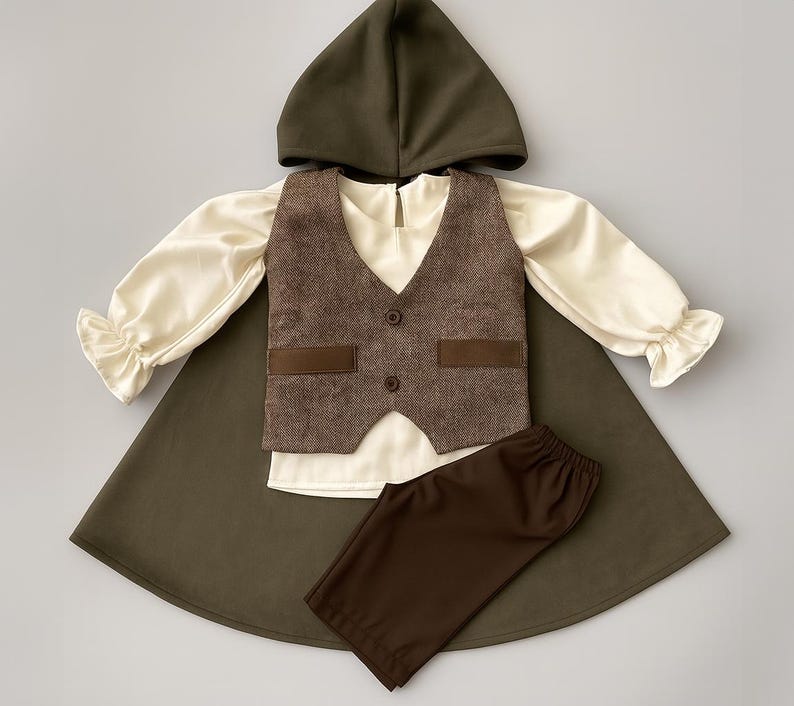 May include: A child's costume set featuring a hooded cape, vest, shirt, and pants. The cape and hood are olive green, the vest is brown tweed, and the shirt is cream-colored with ruffled sleeves. The pants are brown.