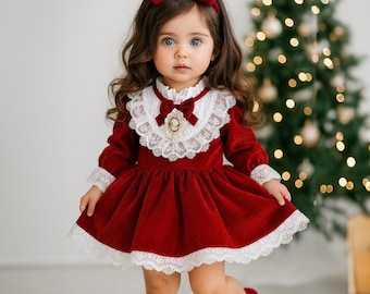 May include: A red velvet dress with white lace trim and a matching red bow headband. The dress features a white lace collar and cuffs, and a decorative brooch. The outfit is completed with red shoes with bows. The background includes a Christmas tree.
