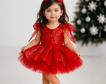 May include: A red sequined dress with a layered tulle skirt and matching red bow headband. The dress features sheer shoulder accents and is paired with red Mary Jane shoes. A Christmas tree is visible in the background.