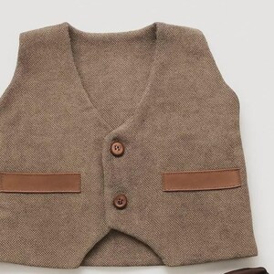 May include: A brown tweed vest with a V-neck, two brown buttons, and two brown leather-like patches. The vest is a classic style, suitable for formal or semi-formal occasions.