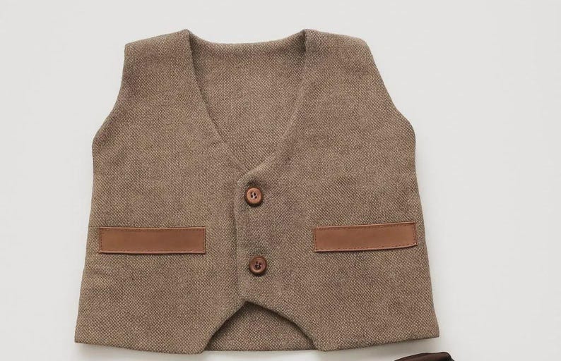 May include: A brown tweed vest with a V-neck, two brown buttons, and two brown leather-like patches. The vest is a classic style, suitable for formal or semi-formal occasions.