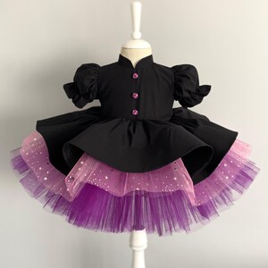 May include: A black dress with a high collar and short, puffed sleeves. The dress features pink buttons and a layered skirt with pink and purple tulle. Gold star accents are visible on the pink layer, creating a whimsical look.