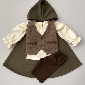 May include: A children's costume set featuring a hooded cape, a cream-colored long-sleeved shirt with ruffled cuffs, a brown tweed vest with two buttons, and brown pants. The cape and hood are olive green.
