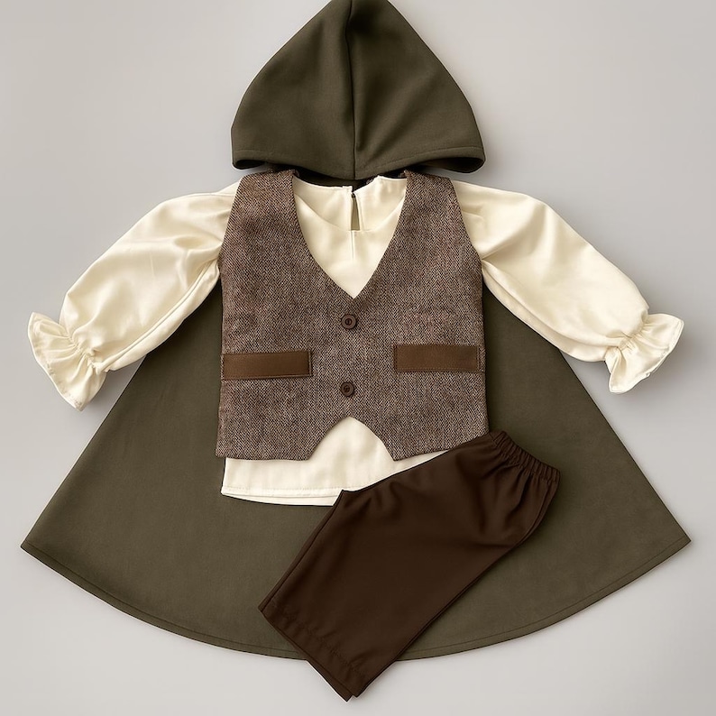May include: A children's costume set featuring a hooded cape, a cream-colored long-sleeved shirt with ruffled cuffs, a brown tweed vest with two buttons, and brown pants. The cape and hood are olive green.