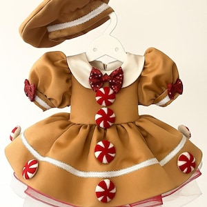 K&ouml;nnte beinhalten: Ein hellbraunes Kleid und eine passende Baskenm&uuml;tze, die einem Lebkuchenkeks &auml;hneln. Das Kleid hat einen wei&szlig;en Kragen, eine rote Schleife und S&uuml;&szlig;igkeitenakzente. Wei&szlig;e Verzierungen und rote Akzente zieren das Kleid und die Baskenm&uuml;tze.