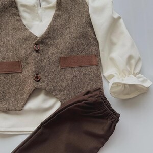 May include: A three-piece outfit for a child, featuring a brown tweed vest with two brown buttons and two brown pockets, a cream-colored long-sleeved shirt, and brown pants. The shirt has ruffled cuffs. The outfit is laid out on a white surface.