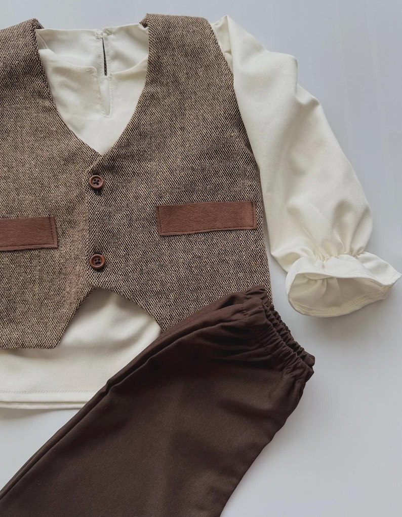 May include: A three-piece outfit for a child, featuring a brown tweed vest with two brown buttons and two brown pockets, a cream-colored long-sleeved shirt, and brown pants. The shirt has ruffled cuffs. The outfit is laid out on a white surface.