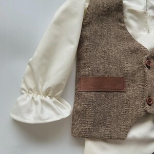May include: A cream-colored long-sleeved shirt with ruffled cuffs, paired with a brown tweed vest featuring a small pocket and wooden buttons. The vest and shirt are part of a formal outfit.