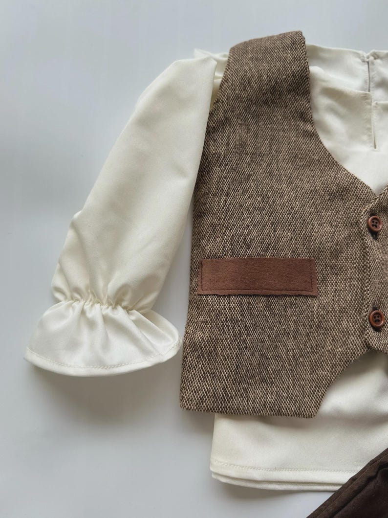 May include: A cream-colored long-sleeved shirt with ruffled cuffs, paired with a brown tweed vest featuring a small pocket and wooden buttons. The vest and shirt are part of a formal outfit.