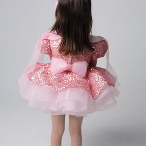 以下が含まれることがあります： ピンクのキラキラしたドレスを着た若い女の子。チュールスカートと背中にピンクのリボンがついています。ドレスは袖が短く、体にフィットしたボディです。