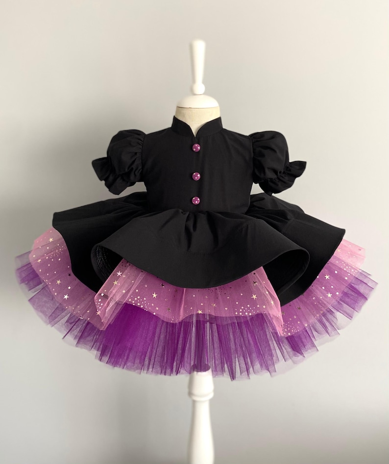 May include: A black dress with a high collar and short, puffed sleeves. The dress features pink buttons and a layered skirt with pink and purple tulle. Gold star accents are visible on the pink layer, creating a whimsical look.