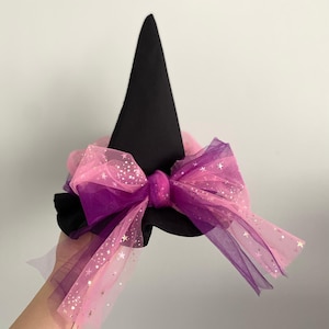 May include: A black witch hat with a large bow. The bow is made of pink and purple tulle, with silver star accents. The hat is a cone shape, and the bow is attached to the base of the hat.