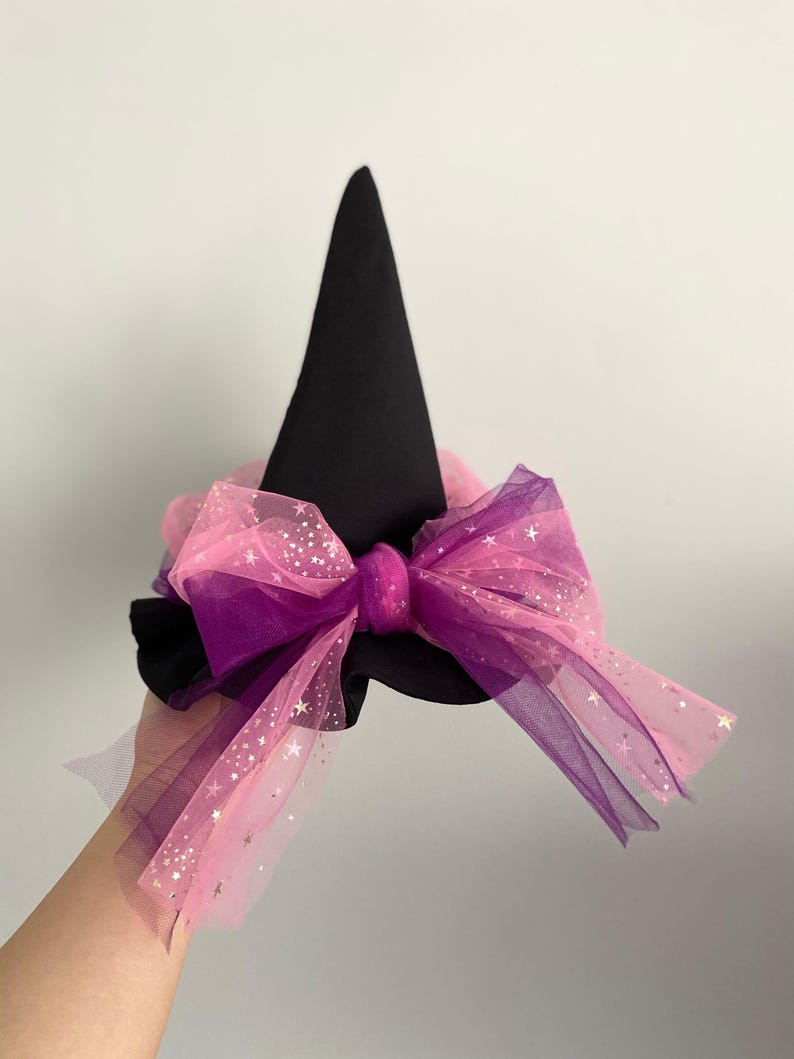 May include: A black witch hat with a large bow. The bow is made of pink and purple tulle, with silver star accents. The hat is a cone shape, and the bow is attached to the base of the hat.