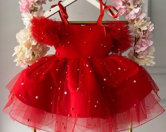 May include: A vibrant red dress featuring a layered tulle skirt and bodice adorned with scattered white pearls. The garment includes off-the-shoulder ruffled tulle sleeves and thin red ribbon shoulder straps, creating a full, voluminous silhouette.