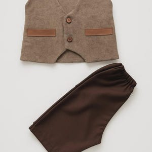 May include: A brown vest and matching pants set. The vest has a textured fabric with two brown leather-like accents and two buttons. The pants are a solid brown color. The set is likely for a child.
