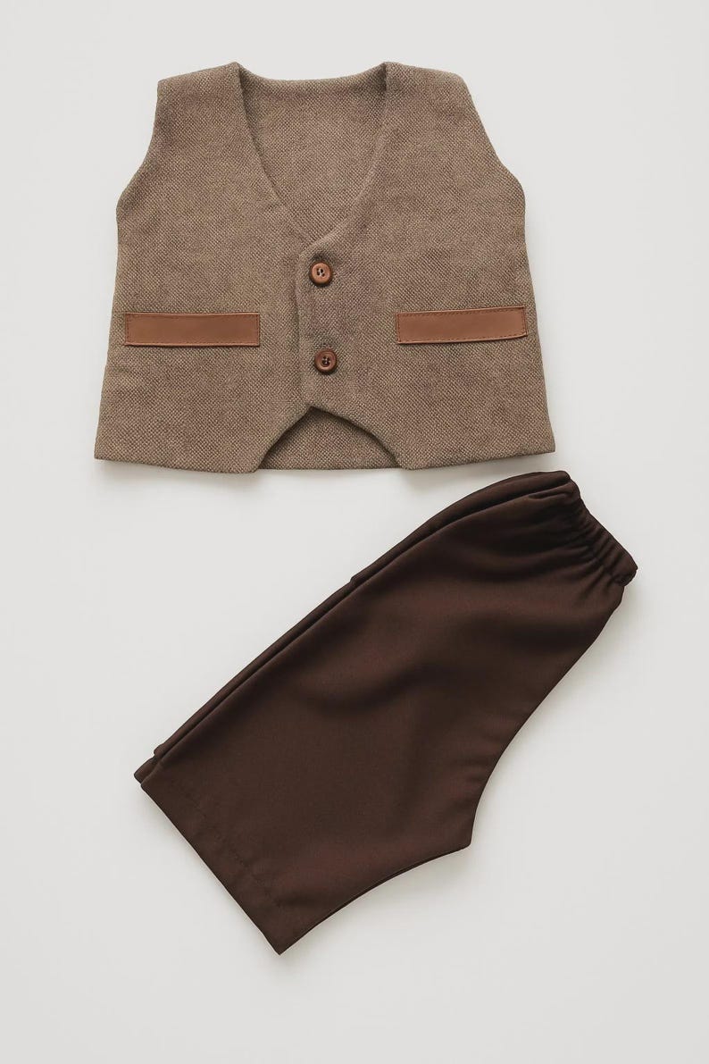 May include: A brown vest and matching pants set. The vest has a textured fabric with two brown leather-like accents and two buttons. The pants are a solid brown color. The set is likely for a child.