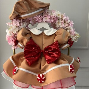 K&ouml;nnte beinhalten: Ein beigefarbenes Kleid mit einer gro&szlig;en roten Pailletten-Schleife, wei&szlig;em Kragen und Puff&auml;rmeln. Das Kleid hat wei&szlig;e Spitzenbes&auml;tze und ist mit roten und wei&szlig;en Bonbon-Akzenten verziert. Ein passender beigefarbener Hut mit wei&szlig;er Bord&uuml;re vervollst&auml;ndigt das Outfit.