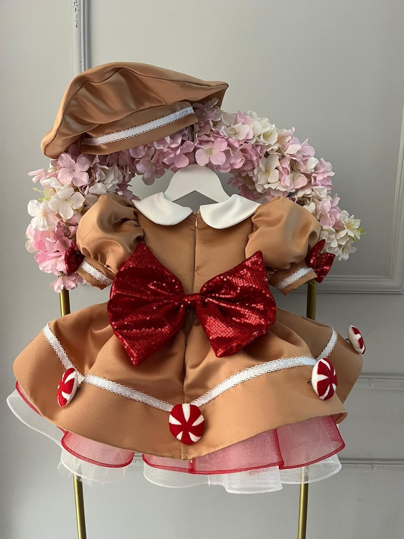 K&ouml;nnte beinhalten: Ein beigefarbenes Kleid mit einer gro&szlig;en roten Pailletten-Schleife, wei&szlig;em Kragen und Puff&auml;rmeln. Das Kleid hat wei&szlig;e Spitzenbes&auml;tze und ist mit roten und wei&szlig;en Bonbon-Akzenten verziert. Ein passender beigefarbener Hut mit wei&szlig;er Bord&uuml;re vervollst&auml;ndigt das Outfit.