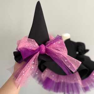 May include: A black witch hat with a large pink and purple bow. The bow is made of layered tulle with star accents. The hat is part of a costume, with a matching skirt visible in the background.