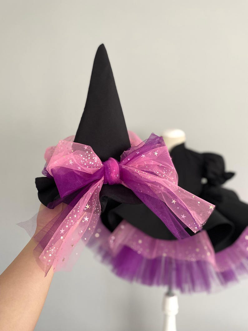 May include: A black witch hat with a large pink and purple bow. The bow is made of layered tulle with star accents. The hat is part of a costume, with a matching skirt visible in the background.