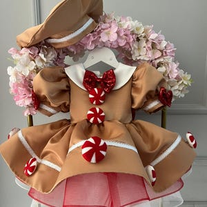 K&ouml;nnte beinhalten: Ein braunes Kleid im Lebkuchen-Thema mit passendem Hut. Das Kleid hat einen wei&szlig;en Kragen, eine rote Schleife und S&uuml;&szlig;igkeiten-Akzente. Der Rock hat eine Unterlage aus rotem und wei&szlig;em T&uuml;ll. Der Hut hat eine wei&szlig;e Bord&uuml;re.