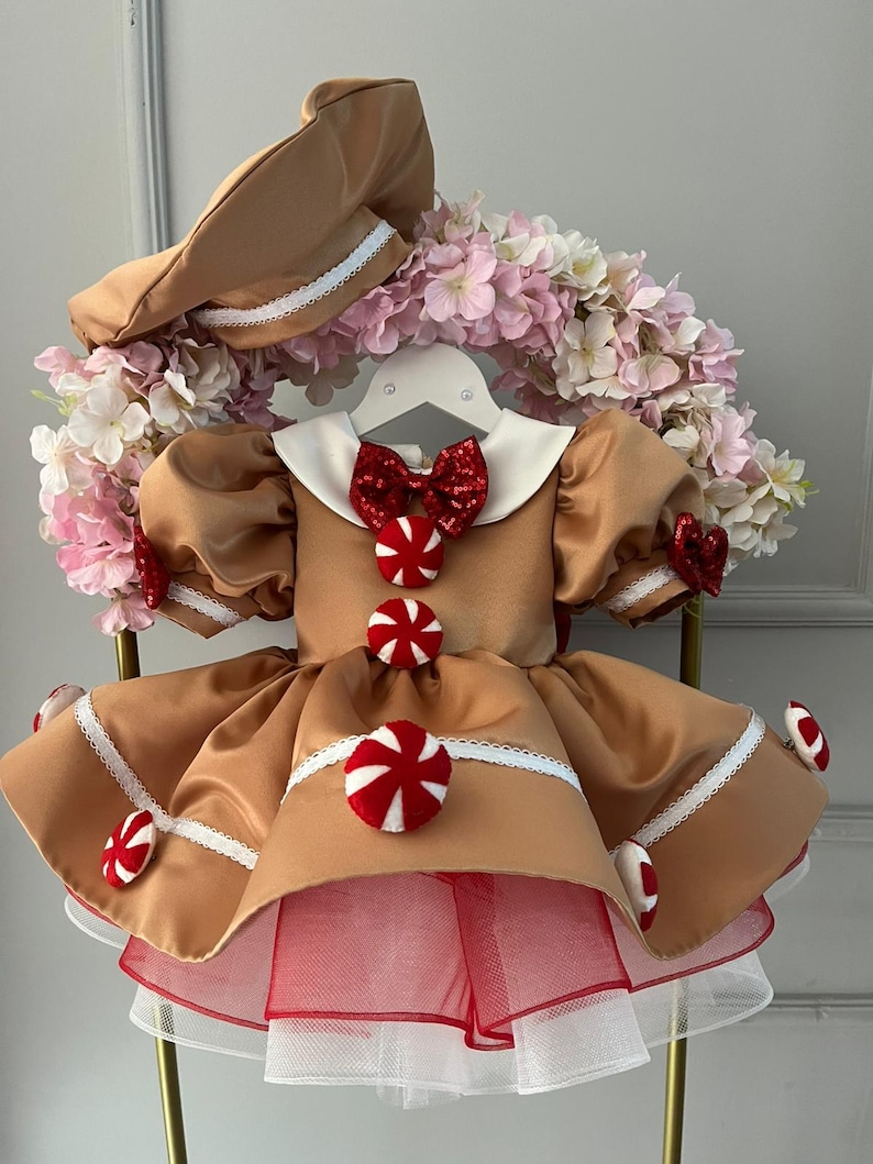 K&ouml;nnte beinhalten: Ein braunes Kleid im Lebkuchen-Thema mit passendem Hut. Das Kleid hat einen wei&szlig;en Kragen, eine rote Schleife und S&uuml;&szlig;igkeiten-Akzente. Der Rock hat eine Unterlage aus rotem und wei&szlig;em T&uuml;ll. Der Hut hat eine wei&szlig;e Bord&uuml;re.