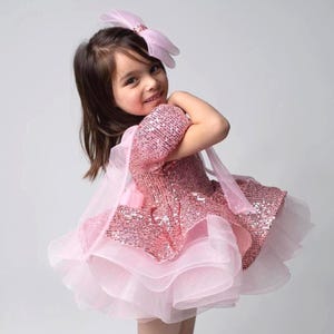 May include: A pink sequined dress with a layered tulle skirt and a matching bow headband. The dress features a short-sleeved bodice and a flowing skirt. The overall design is a light pink color, perfect for a special occasion.
