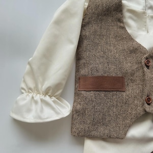 May include: A cream-colored long-sleeved shirt with ruffled cuffs, paired with a brown tweed vest featuring a small pocket and wooden buttons. The vest and shirt are part of a formal outfit.
