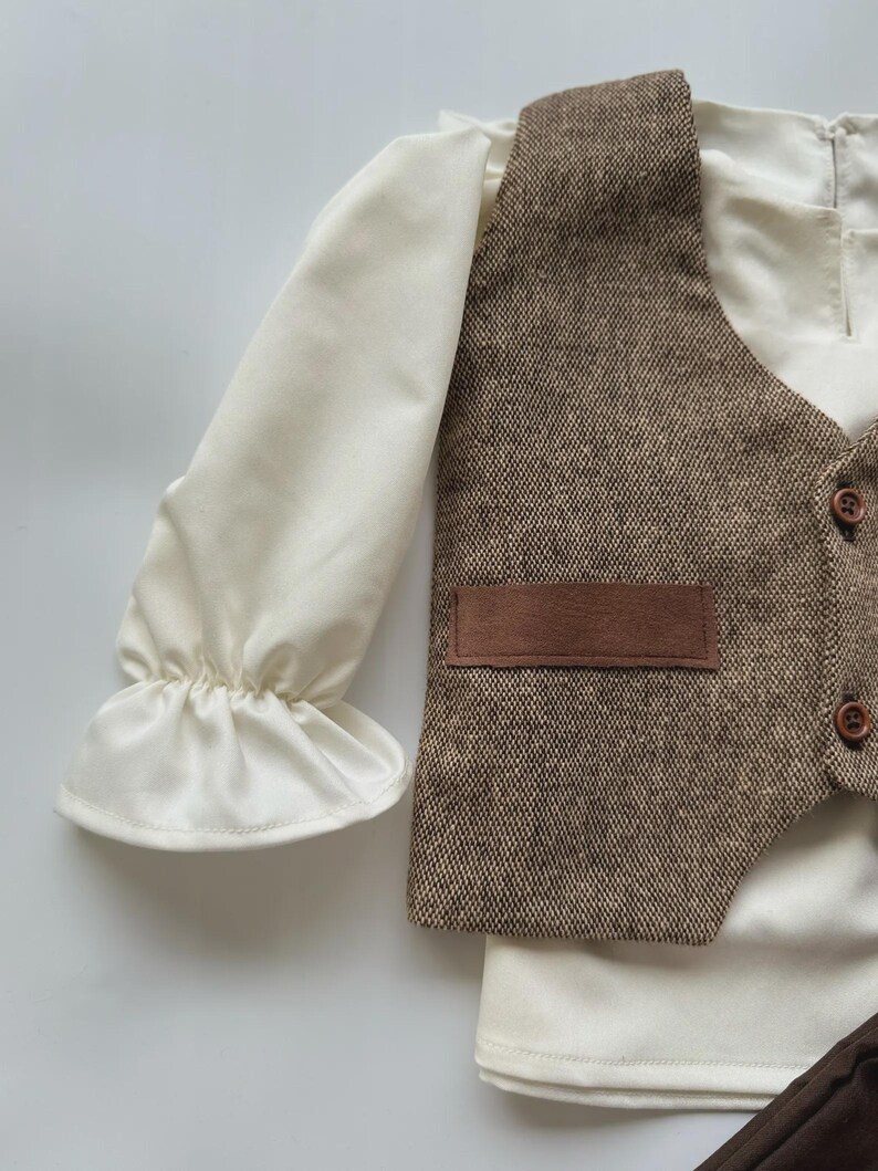 May include: A cream-colored long-sleeved shirt with ruffled cuffs, paired with a brown tweed vest featuring a small pocket and wooden buttons. The vest and shirt are part of a formal outfit.