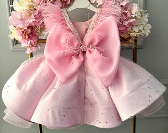 May include: A light pink dress with a large bow and feather accents. The dress features a V-neckline adorned with pearls and a full skirt with silver sequins. The dress is displayed on a gold hanger against a backdrop of pink flowers.