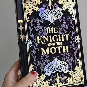 May include: A hardback book titled "The Knight and the Moth" with a black cover and gold foil detailing. The cover design includes a moth illustration and floral patterns. A decorative bookmark is visible.
