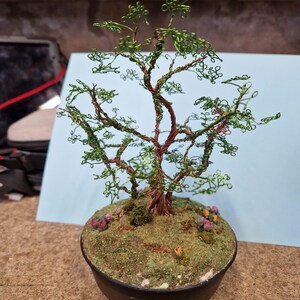 May include: A wire sculpture of a tree with green leaves and a brown trunk. The tree is in a black pot with green moss and pink flowers.