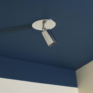 Könnte beinhalten: Ein moderner, silberfarbener Deckenstrahler. Das Licht ist auf einer kreisförmigen Basis montiert und nach unten abgewinkelt. Der Hintergrund ist eine tiefblaue Decke und eine weiße Wand.