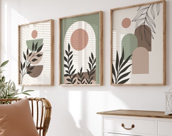 May include: Three framed abstract art prints with a minimalist design featuring geometric shapes, leaves, and a circle in shades of green, brown, and pink. The prints are hanging on a white wall above a white cabinet with a wooden top.