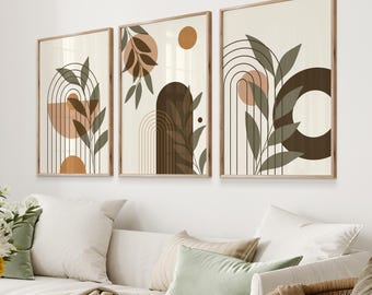 May include: Three framed art prints with abstract botanical designs. Each print features a beige background with brown, orange, and green leaf and arch motifs. The frames are a light brown colour, and the prints are displayed above a white sofa.